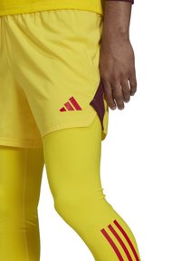 Gele sportshorts met rode accenten en branding, gecombineerd met gele compressieleggings met rode strepen aan de zijkant.