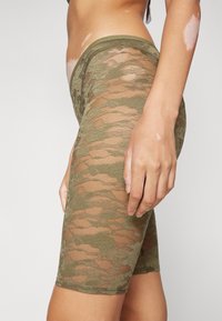 Calças ciclismo em malha verde oliva com padrão de camuflagem, cintura elástica e detalhe texturizado no tecido.