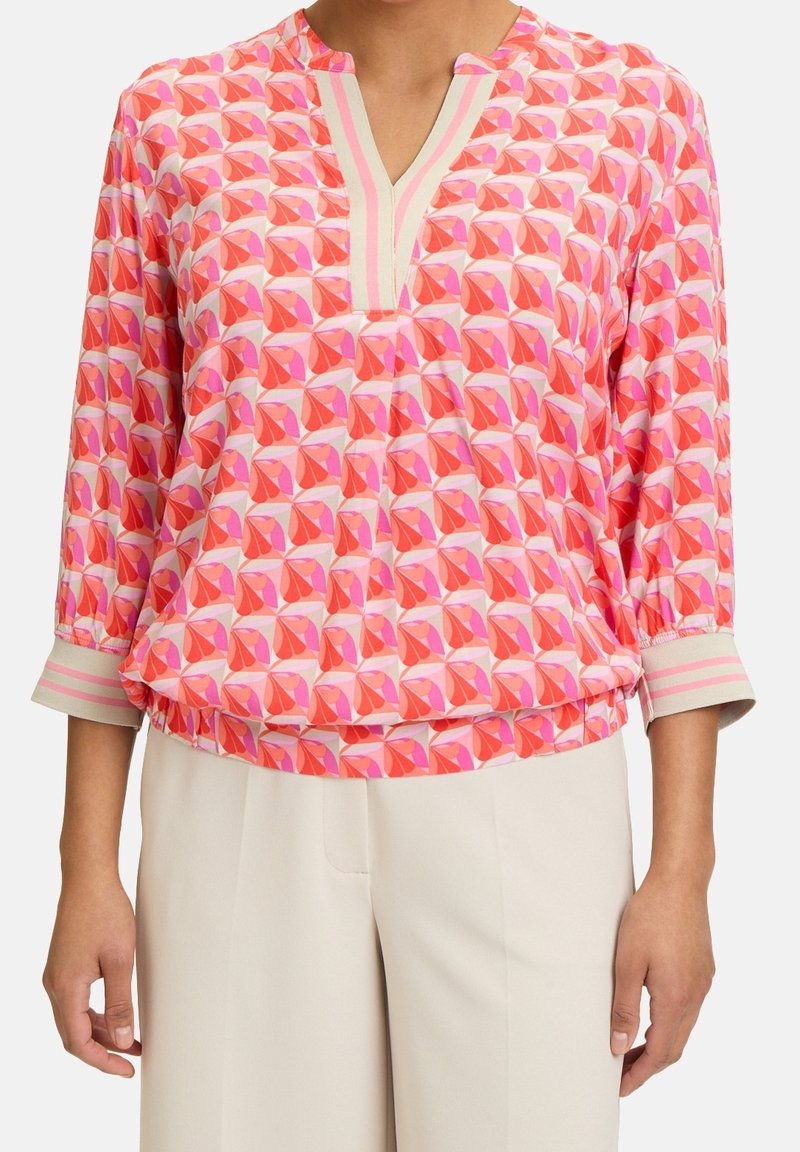 Femme portant un chemisier à motifs géométriques rose, rouge et orange avec des manches trois-quarts et un pantalon blanc.