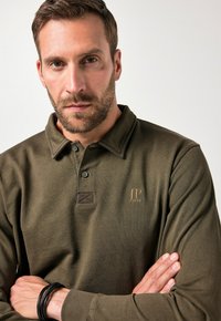 Oliwkowa koszula polo z długim rękawem, z kołnierzykiem zapinanym na guziki, teksturowana tkanina oraz haftowany logo na piersi. Wyróżniający się szczegół to zapięcie na guziki.