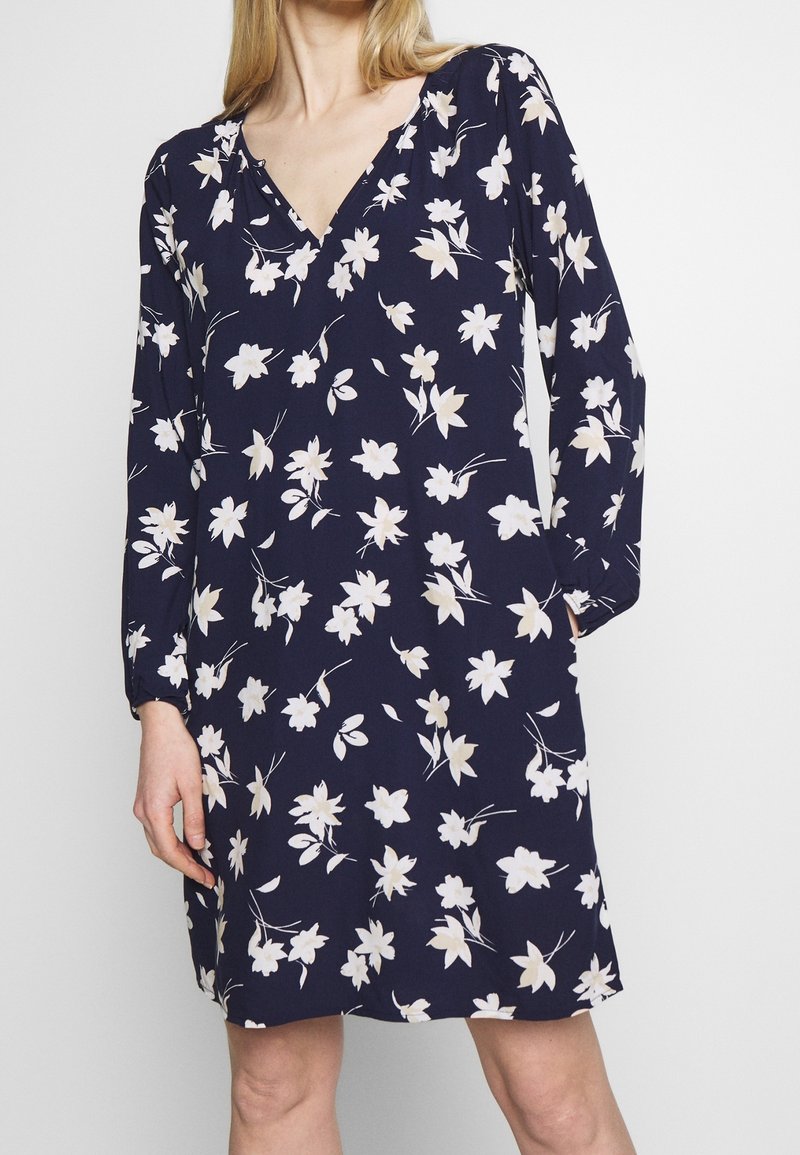 Femme portant une robe bleu marine à manches longues avec un imprimé floral blanc et un col en V, debout devant un fond blanc uni.