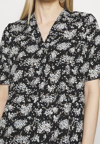 Blouse florale noire à manches courtes, avec des motifs floraux blancs et bleu pâle, un décolleté en V et une coupe ample. Texture de tissu lisse.