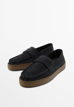 Paar schwarze Wildleder-Slipper mit breitem Riemendetail und braunen Gummisohlen auf weißem Hintergrund.