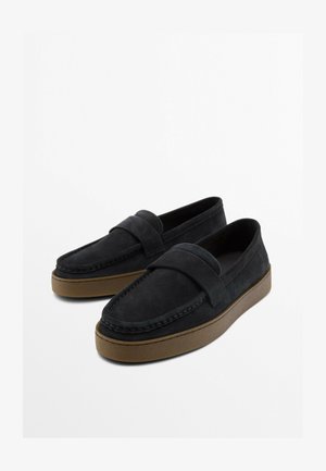 Paire de mocassins en suède noir avec une large bride et des semelles en caoutchouc marron sur un fond blanc uni.