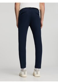 Pantaloni slim-fit blu navy con una texture liscia, dotati di tasche posteriori e una vita standard, abbinati a sneakers bianche.
