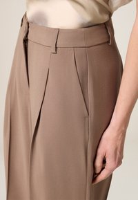 Beige, maßgeschneiderte Hose mit Falten, ausgestattet mit einem Reißverschluss und seitlichen Taschen. Der Stoff hat eine glatte, strukturierte Oberfläche.