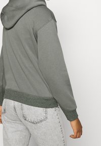Felpa grigia con cappuccio e orli a coste, caratterizzata da una texture morbida. Indossata con jeans in denim grigio chiaro.