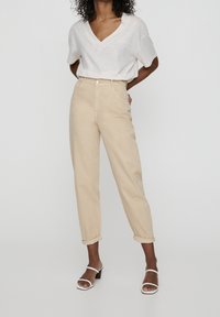 Osoba ubrana w beżowe spodnie z wysokim stanem i podwiniętymi nogawkami, białą bluzkę z dekoltem w serek i krótkim rękawem oraz białe sandały na obcasie, stojąca na tle jednolitej ściany.