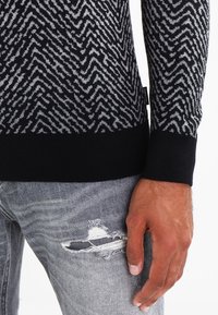 Schwarz gestrickter Pullover mit grauem Zickzackmuster; gerippte Bündchen; kombiniert mit zerrissenen grauen Jeans mit zerfetzten Stoffakzenten.