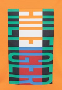 Oranžové tričko s multicolor grafickým designem, na kterém se nacházejí tučné, překrývající se písmena v zelené, černé, modré, červené a bílé barvě.