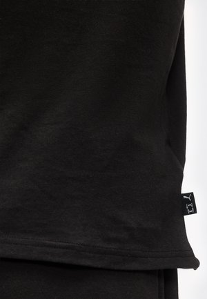 Fekete szövet látható szegélyvarrással és egy kis fekete címkével az oldalsó varráson, amelyen fehér Puma és Under Armour logók láthatók.