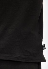 Μαύρο ύφασμα με ορατή ραφή στο τελείωμα και μια μικρή μαύρη ετικέτα στη ραφή της πλευράς, που φέρει λευκά λογότυπα Puma και Under Armour.