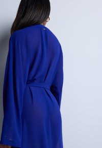 Robe en satin bleu profond à manches longues, avec une taille ceinturée, arborant un subtil éclat et des détails en doré au niveau du col. Texture lisse.