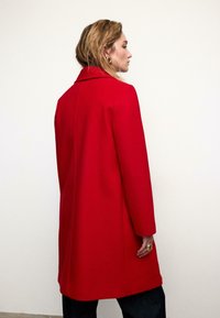 Rød uldfrakke med klassisk krave, lange ærmer og en afslappet pasform. Bemærkelsesværdigt sømløst design og minimalistisk stil uden synlige lukninger.