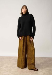 Czarny sweter w robiony na drutach z wysokim kołnierzem, zestawiony z szerokimi, prążkowanymi spodniami w musztardowym kolorze oraz ciemnobrązowym obuwiem. Prosty, teksturowany wzór.