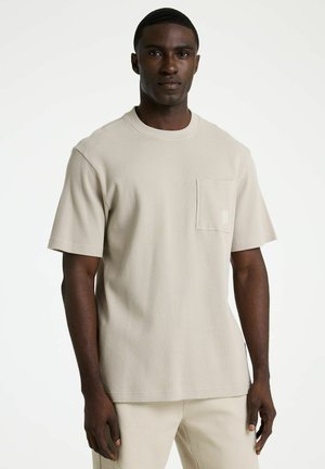 Man die een beige textuur kort mouwen overhemd met borstzak en bijpassende broek draagt, staand tegen een witte achtergrond.