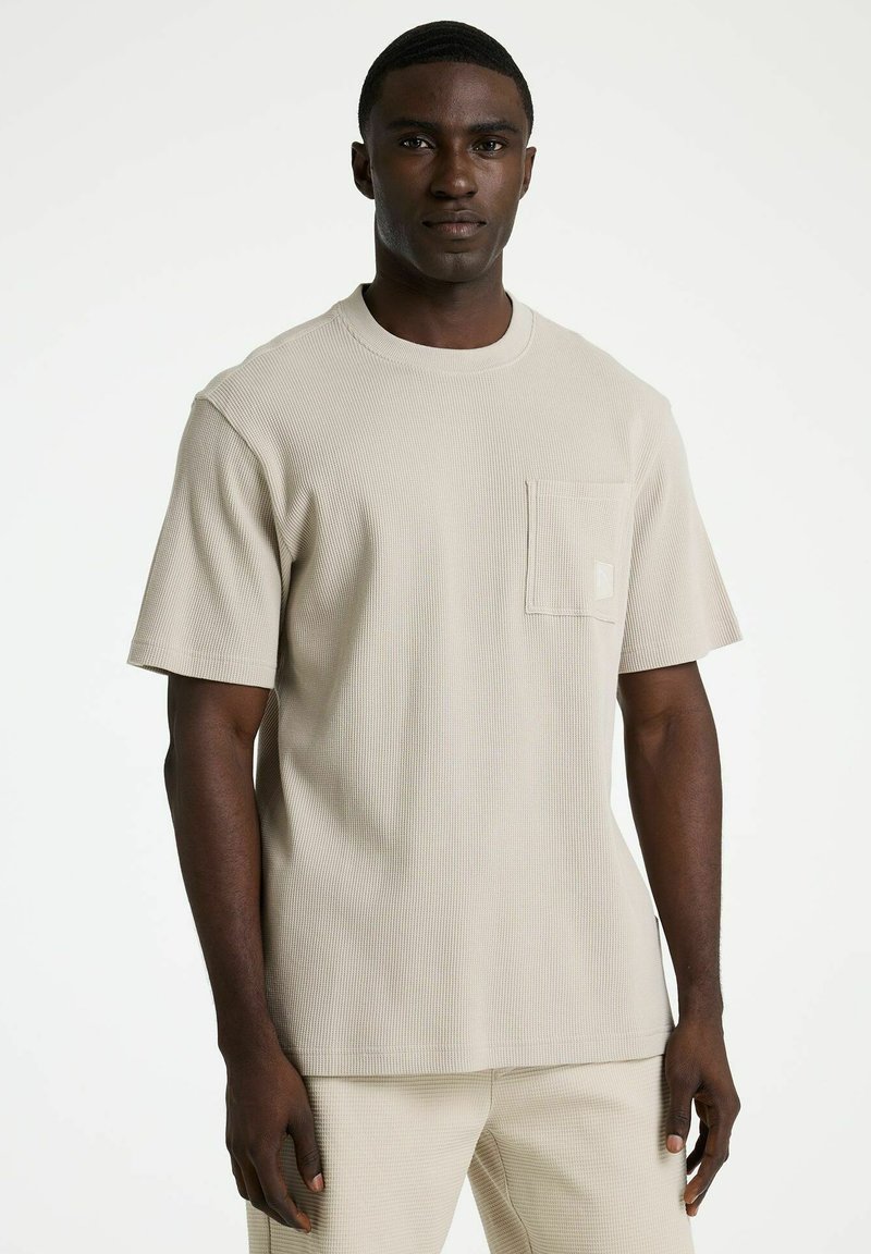 Man die een beige textuur kort mouwen overhemd met borstzak en bijpassende broek draagt, staand tegen een witte achtergrond.