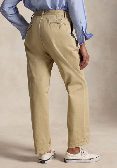 Persoon die beige broek, lichtblauw shirt en witte sneakers draagt, met één hand bij de zij, gezien van achteren.