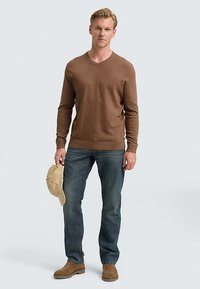 Bruine gebreide V-hals trui, blauwe spijkerbroek en bruine suède enkellaarsjes. Model houdt een lichtgekleurde pet vast. Neutrale achtergrond.