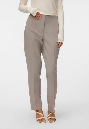 Femme portant un pantalon beige taille haute ajusté, associé à des sandales blanches ouvertes aux orteils et un haut crème à manches longues, debout devant un fond blanc.