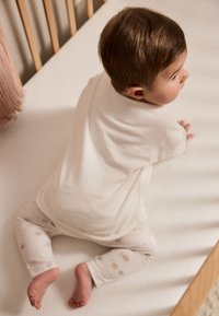Camiseta de algodón blanca, pantalones suaves con puntos multicolores, niño sentado en un colchón cubierto con una tela clara, marco de cuna de madera visible.