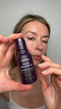 Woman holding a small dark purple bottle of Aveda Invati Ultra Advanced exfoliating shampoo near her face against a plain background.