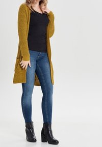 Dlouhý hořčicový cardigan s žebrovanou texturou, černé tílko a modré úzké džíny. Černé kotníkové boty na podpatku. Jednoduchý, funkční design.