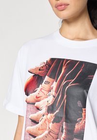 Bílý bavlněný tričko s velkým grafikou obuvi na basketbalovém hřišti, zobrazující světlé barvy a podrobné designové prvky.