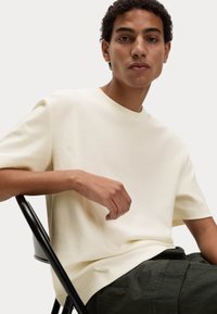 Korte mouwen, textuur crème t-shirt van gebreide stof, gecombineerd met donkergroene broek, model zittend op een zwarte stoel.