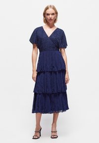 Vestido azul marino en capas con detalles de cuentas. Escote en forma de V, mangas cortas con vuelo, tejido fluido y volantes superpuestos para dar textura y movimiento.