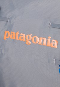 Γκρίζο ύφασμα με λεία υφή φέρει τη λέξη "patagonia" σε ανυψωμένα πορτοκαλί γράμματα. Περιλαμβάνει μπλε λεπτομέρειες και ραφές.