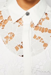Chemise en dentelle blanche avec motifs floraux, col pointu et une seule boutonnière au col laissant apparaître la peau en dessous.