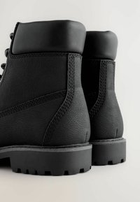 Schwarze Lederknöchelstiefel mit gepolsterten Kragen, detaillierten Nähten und dicken Profilsohlen, von hinten auf einem hellen Hintergrund gezeigt.