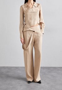 Beige Zweiteiler bestehend aus einem gekürzten, asymmetrischen Langarmoberteil mit Knopfdetails und hoch taillierten, weiten Hosen. Schwarze Schuhe.
