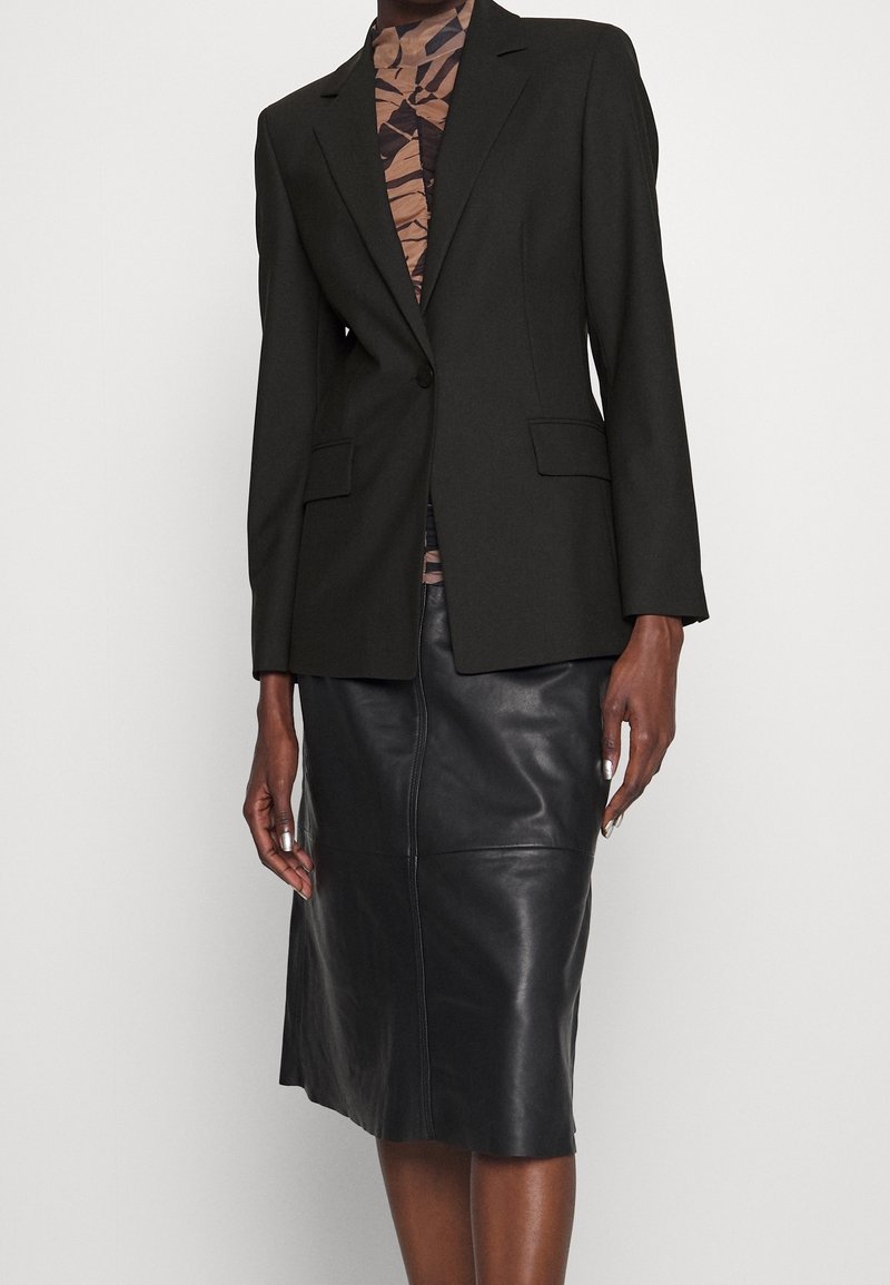 Blazer noir sur un haut marron et noir à motifs, associé à une jupe en cuir noire longueur genou sur une personne debout.