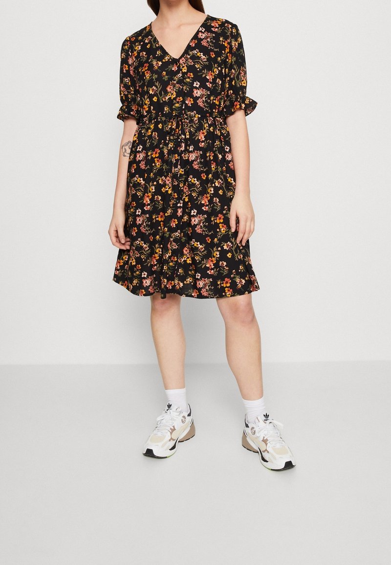 Femme portant une robe noire à fleurs mi-longue avec des manches bouffantes et des baskets blanches, debout devant un fond gris clair uni.