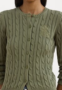 Cardigan en tricot torsadé vert avec un col rond, des boutons devant et des poignets côtelés. Présente un logo brodé sur le côté gauche.