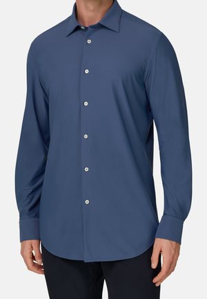 Homme portant une chemise bleue unie à manches longues boutonnée avec des poignets boutonnés et un pantalon noir sur un fond uni.
