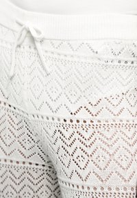 Pantalon en maille blanche avec taille à cordon, présentant un motif en dentelle géométrique détaillé et des sections semi-transparentes.