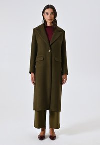 Cappotto lungo verde oliva con colletto a vista, bottone singolo, due tasche anteriori e tessuto strutturato, abbinato a pantaloni coordinati e scarpe borgogna.
