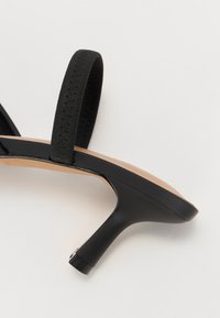 Must mustaga sussud musta tekstureeritud rihmaga, silebeige jalanõu ja peen must konts. Omab minimalistlikku disaini ja avatud varbaosa.