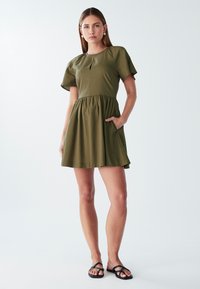 Olivno zelena obleka s kratkimi rokavi in okroglo ovratnikom, podrobnostjo v obliki ključavnice, nabranim pasom in stranskima žepoma. Gladka tkanina z udobnim krojem.