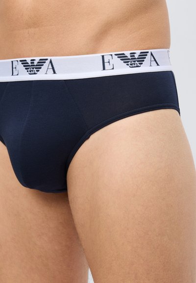 Inimese lähivõte, kes kannab tumelisi siniseid aluspesu valge vöökohaga, millel on mustad "EA" ja kotkasteemalised logod.