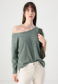 Grønn strikkegenser med off-the-shoulder design, teksturert stoff, lange ermer og løs passform. Brukt sammen med lyse jeans og en veske.