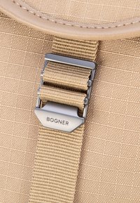 Close-up bežnega platnenega nahrbtnika s svetlo rjavim trakom, ki ga drži srebrna kovinska zaponka, vgravirana z "BOGNER."