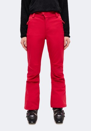 Une personne portant un pantalon de ski rouge vif, un haut noir et des chaussures de ski noires, debout devant un fond clair et uni.