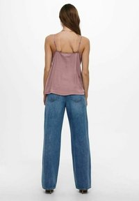 En modell står med ryggen mot kameran, iklädd en åtsittande mauve linne och lösa blå jeans framför en plain vit bakgrund.