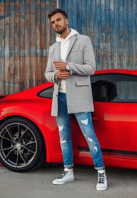 Cappotto grigio su misura sopra una felpa bianca, abbinato a jeans blu strappati e sneaker alte bianche, accanto a una macchina sportiva rossa.