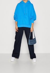 Felpa blu in cotone con tasca frontale, abbinata a jeans neri a gamba larga e sneaker bianche con piattaforma. Viene tenuto in mano una piccola borsa a spalla in denim.