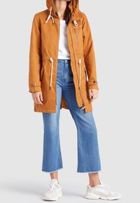 Pomarańczowa wodoodporna kurtka z kapturem, sznurkami, zamkiem z przodu i patkowymi kieszeniami, zestawiona z niebieskimi skróconymi dżinsami i białymi sneakersami.